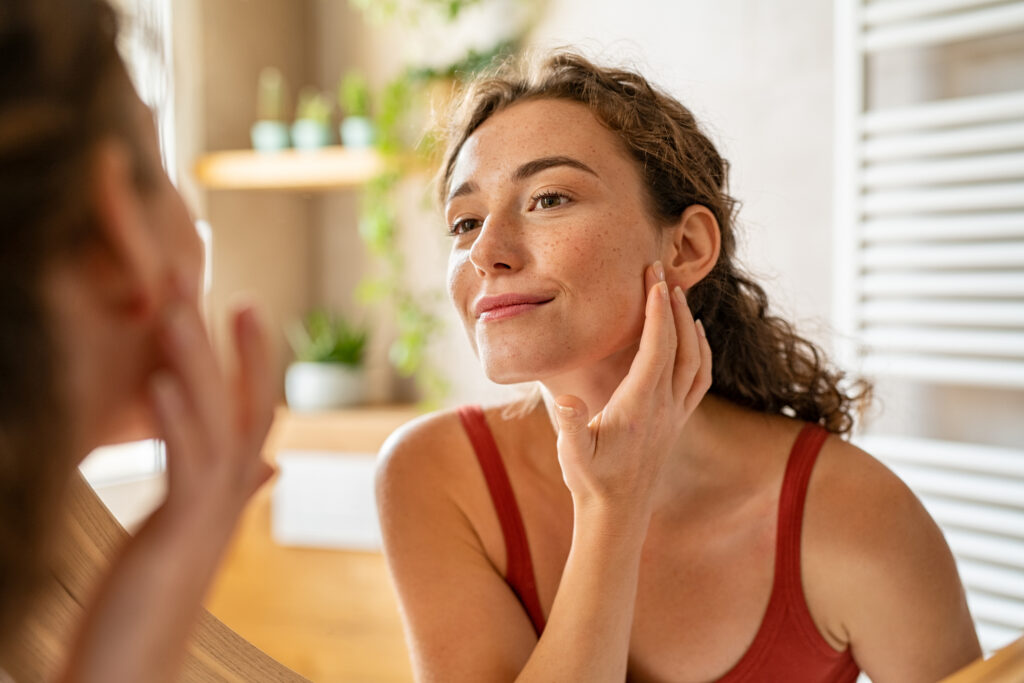 Woman examining her skin and applying moisturizer as part of Acne Bootcamp skincare routine at Reimagine Beauty Lancaster Med Spa.